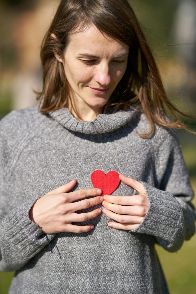 pexels-photo-1820509-1820509 A woman outdoors holding a red heart against her chest, capturing a warm moment.