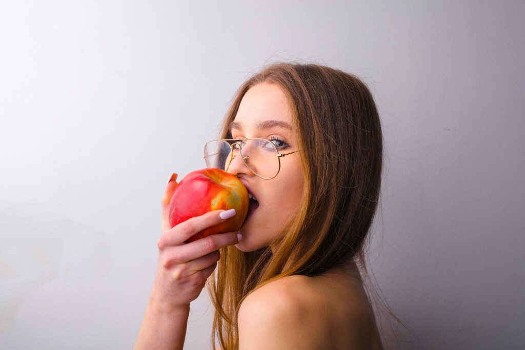 pexels-photo-3765117-3765117 A young woman in glasses takes a bite of a fresh red apple against a gray wall.