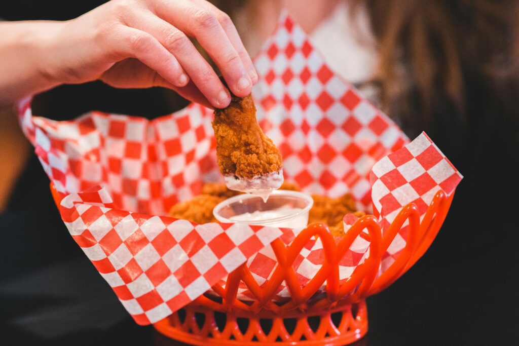 Hand dips crispy fried chicken into creamy sauce in a checkered basket.