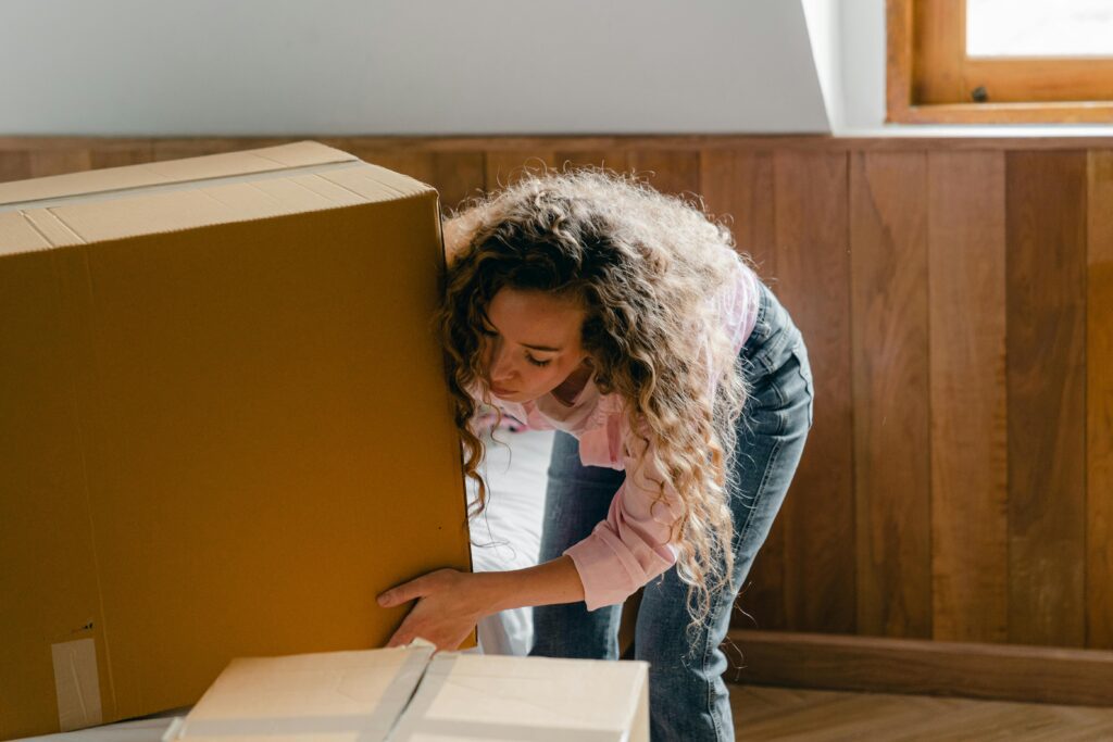 pexels-photo-4246065-4246065 Woman carrying a large cardboard box in a bright, modern interior, signifying moving or relocation.