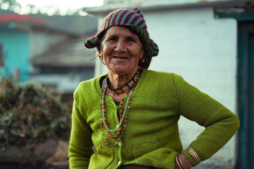 A joyful elderly woman wearing a green knitted sweater and colorful beanie smiles warmly outdoors.