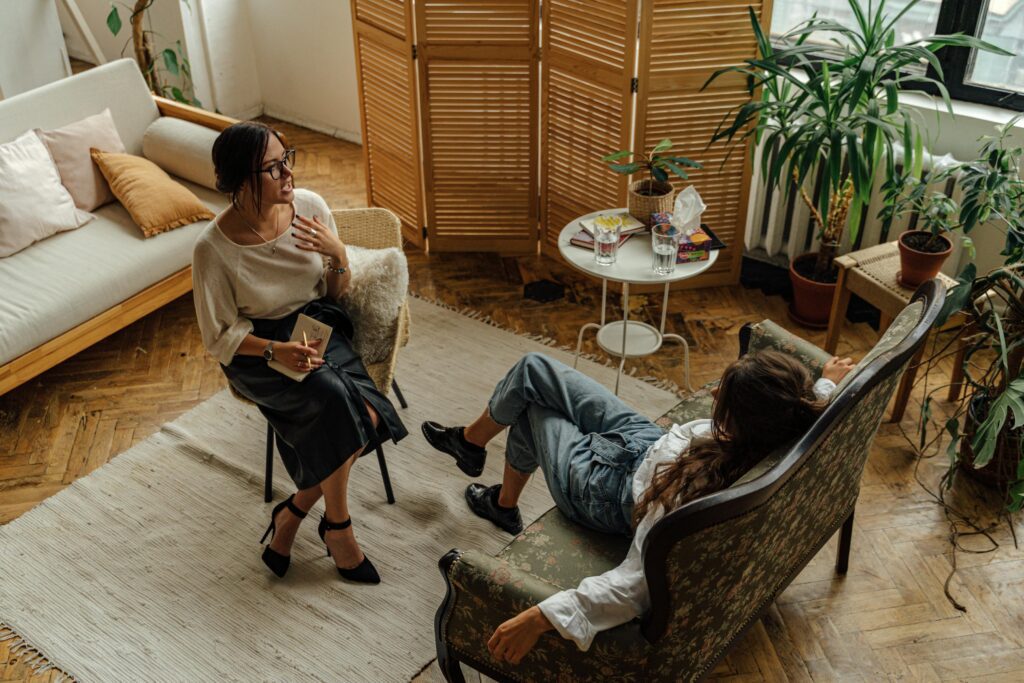 pexels-photo-5217852-5217852 Two women engaged in a psychotherapy session in a warm, inviting interior with plants and natural lighting.