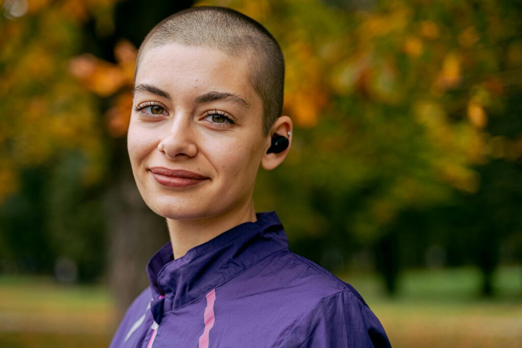 pexels-photo-5588323-5588323 Portrait of a woman with a buzzcut wearing earbuds outdoors in autumn.