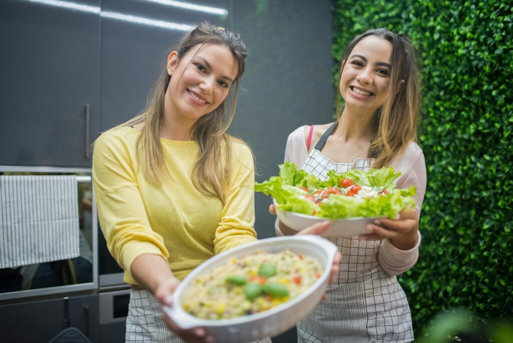 pexels-photo-7964687-7964687 Two women joyfully present fresh vegetable salads in a modern kitchen setting, emphasizing healthy eating.