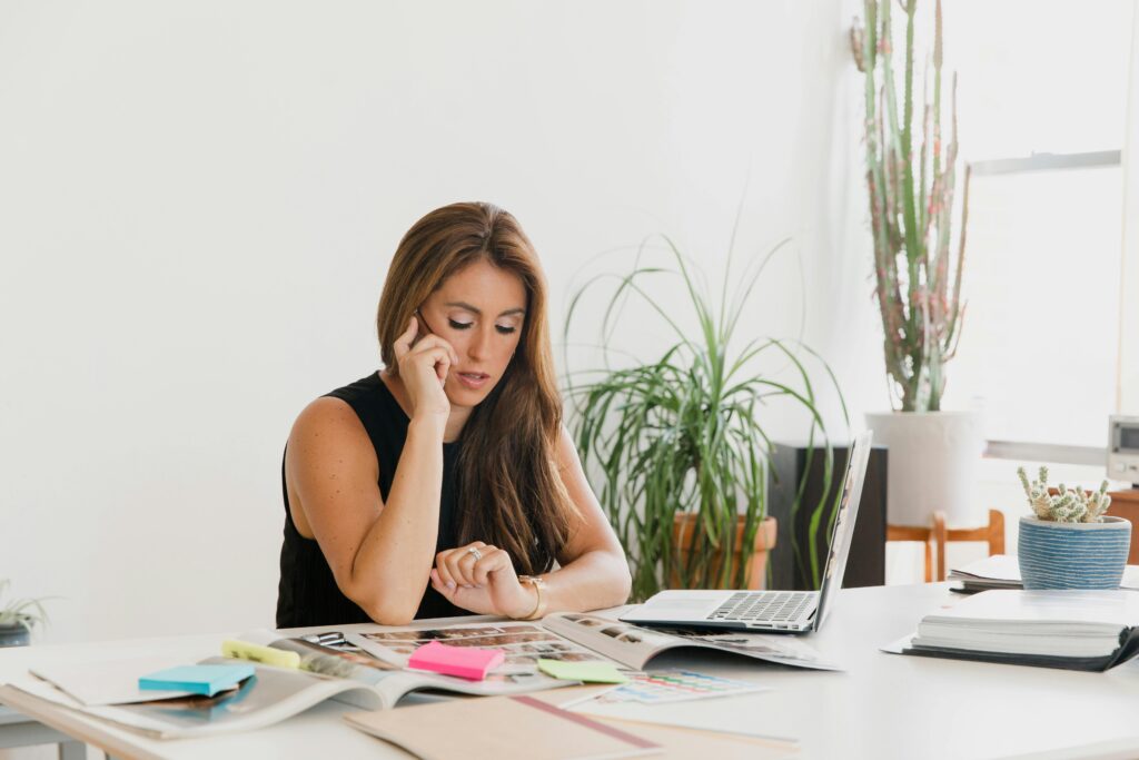 Confident woman multitasking with phone and laptop in a bright, plant-filled office space.