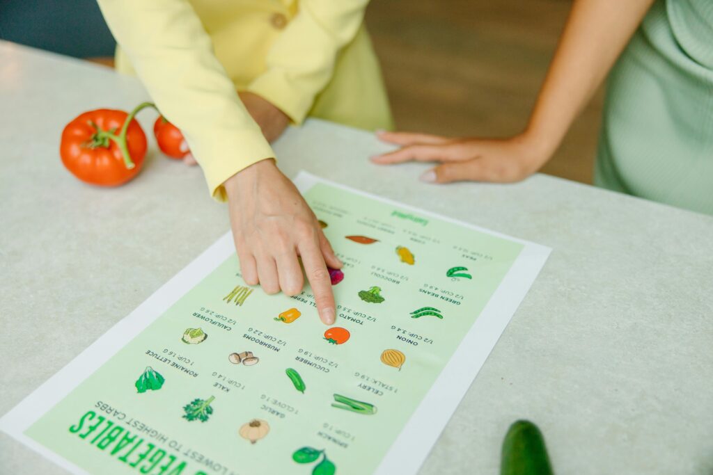 pexels-photo-8844387-8844387 Close-up of hands pointing to a vegetable nutrition chart with fresh tomatoes on the table.