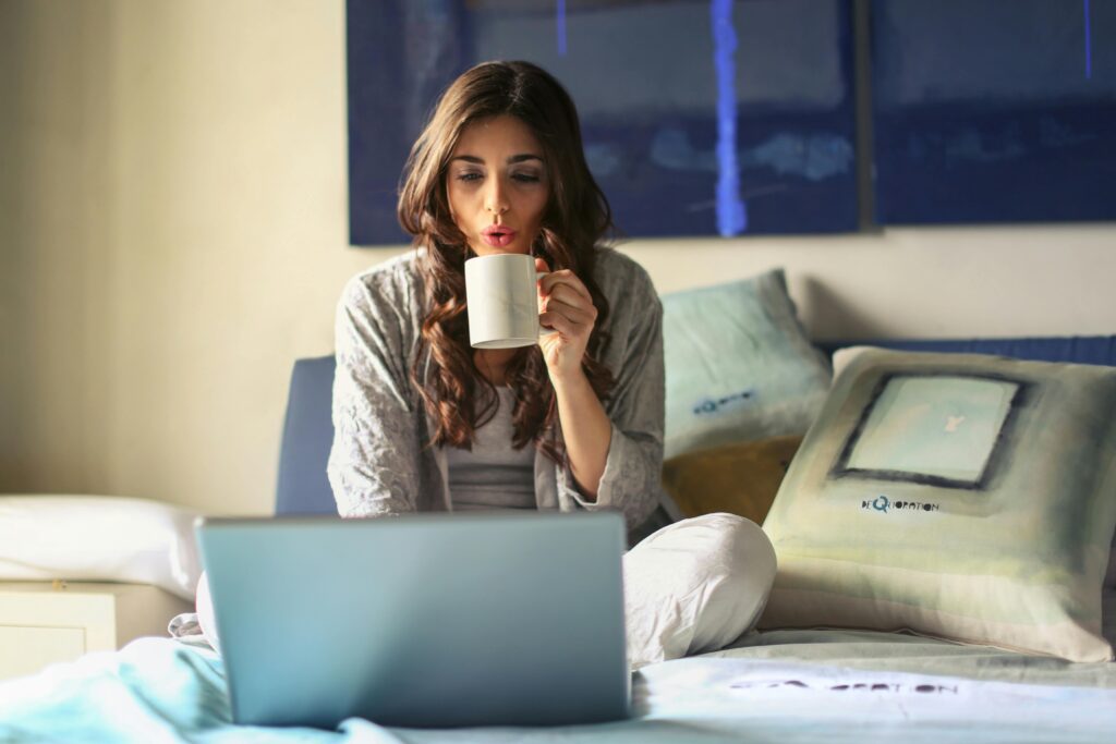 pexels-photo-935743-935743 A woman enjoying coffee while working from home in a cozy bedroom setting with a laptop.