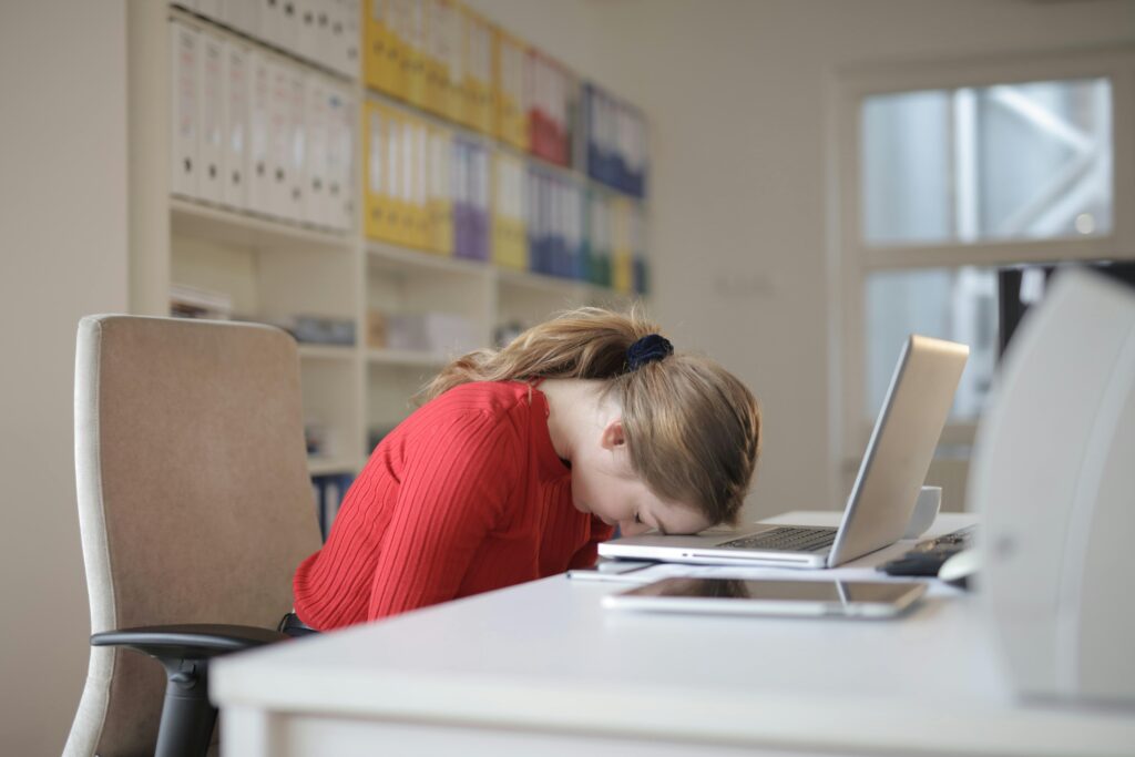 pexels-photo-3791136-3791136 A tired woman in a red sweater leans her head on a desk with a laptop, symbolizing workplace fatigue.