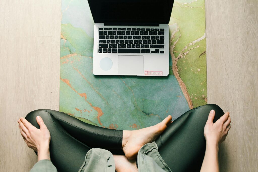 Adult sitting cross-legged on a yoga mat using a laptop, viewed from above.