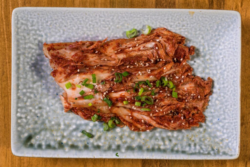 pexels-photo-5774156-5774156 Close-up of fresh homemade kimchi garnished with green onions and sesame seeds on a textured plate.